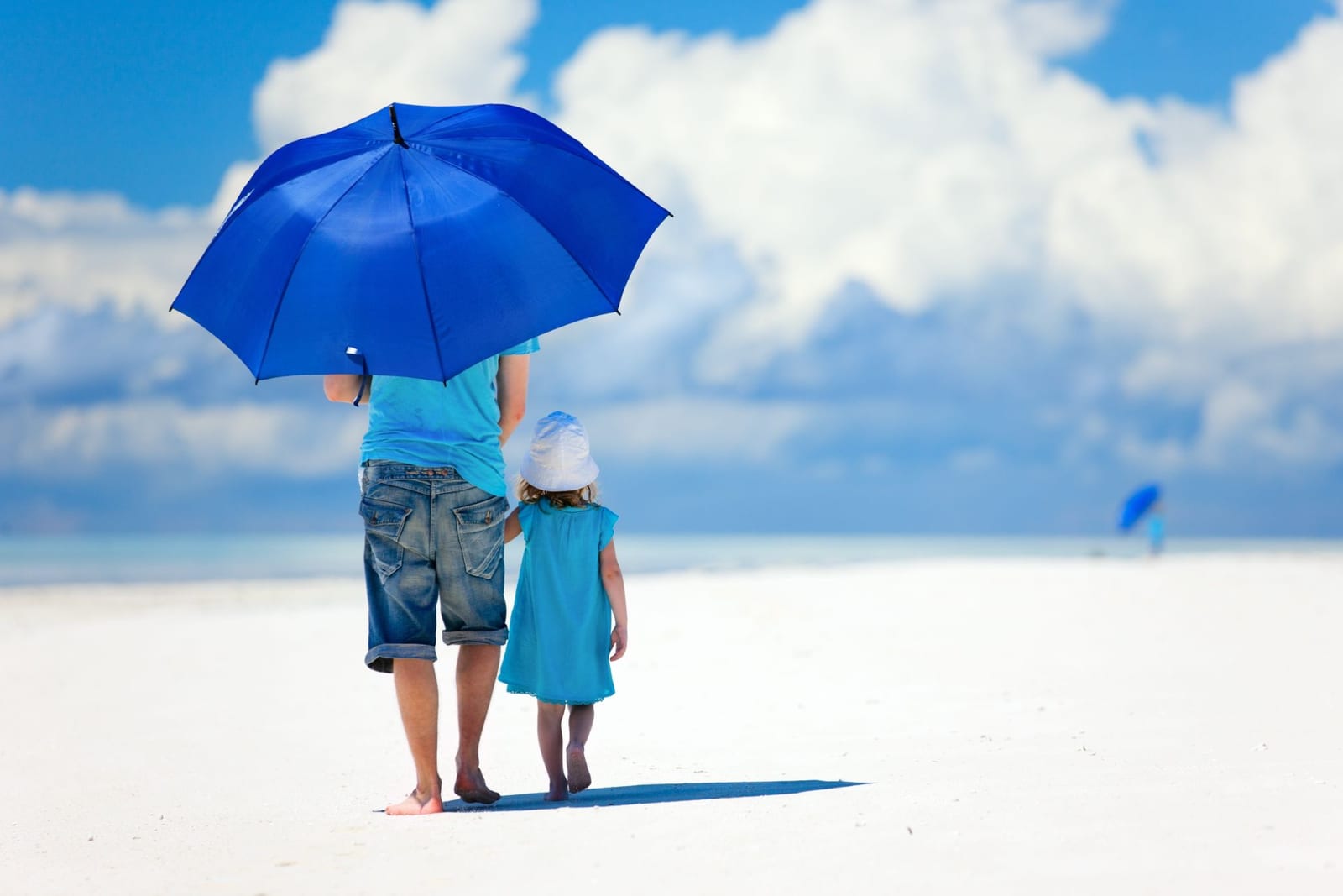 Una madre e una bambina camminano tenendosi per mano su una spiaggia, riparate da un ombrello blu